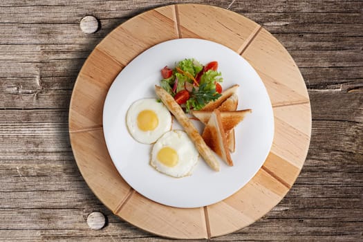 Top view of fried eggs with homemade sausage, toasts and fresh tomato and salad on wooden table.