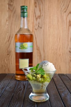 A cup of apple ice cream decorated with kiwi, mint leaves and poppy. Bottle of syrup on background.
