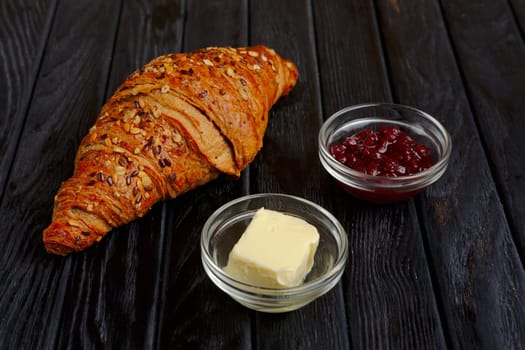 Croissant with jam and butter on dark wooden table.