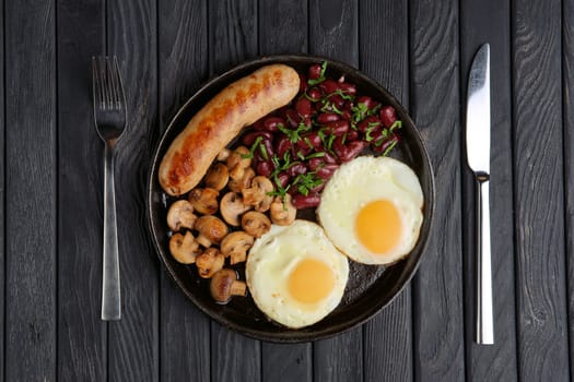 Cast-iron frying pan with fried eggs, sausage, mushroom and beans on wooden table. Top view.