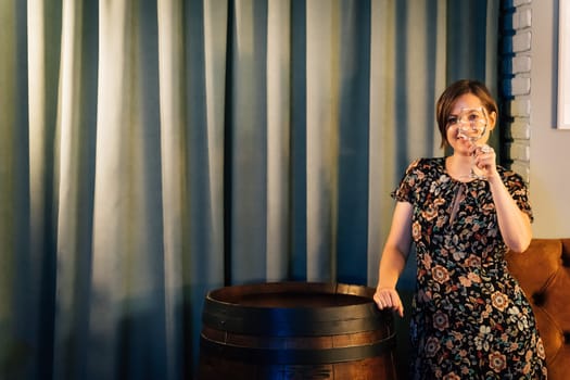 A woman closes her eye with a wine glass standing near a wine barrel.