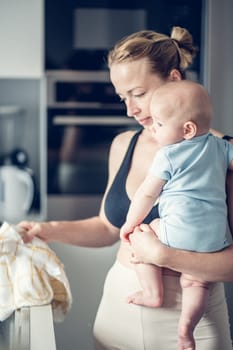 Woman wiping kitchen sink with a cloth after finishing washing the dishes while holding four months old baby boy in her hands.