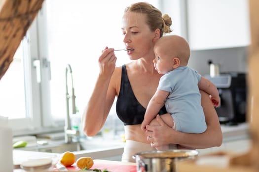 Woman cooking and tasing food while holding four months old baby boy in her hands.