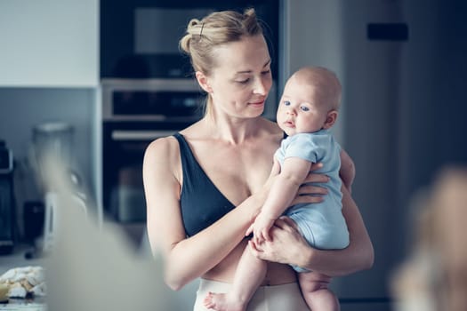 Pretty young mother holding her newborn baby boy standing near kitchen window at home.