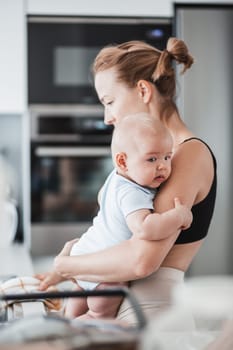 Woman wiping kitchen sink with a cloth after finishing washing the dishes while holding four months old baby boy in her hands.