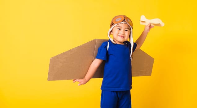 Happy Asian handsome funny child or kid little boy smile wear pilot hat play and goggles with toy cardboard airplane wings fly hold plane toy, studio shot isolated yellow background, Startup freedom