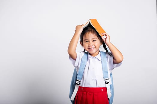 Asian adorable toddler smiling happy wearing student thai uniform red skirt stand holding book over head like roof in studio shot isolated on white background, Portrait little children girl preschool