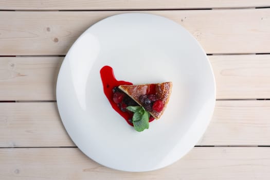 Top view of cheesecake with berries, sauce and green mint leaf on plate on wooden table.