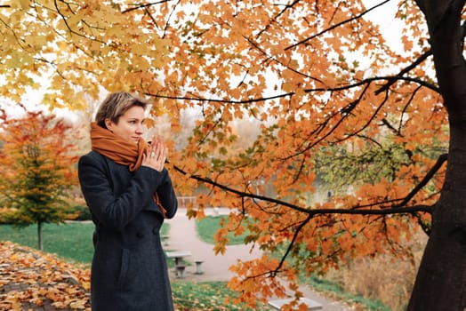Short-haired blonde in coat and scarf warms her hands in the park.