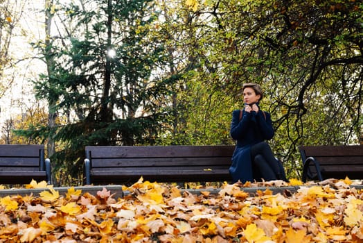 A woman wraps herself in a coat sitting on a bench in an autumn park.