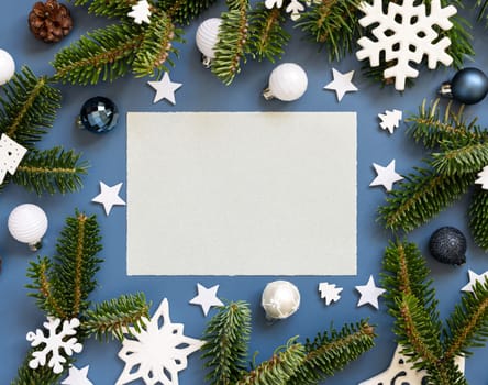 Blank paper card on a blue background near white Christmas decorations and fir branches top view, card mockup, copy space.. New Year and Christmas holiday atmospheric flat lay
