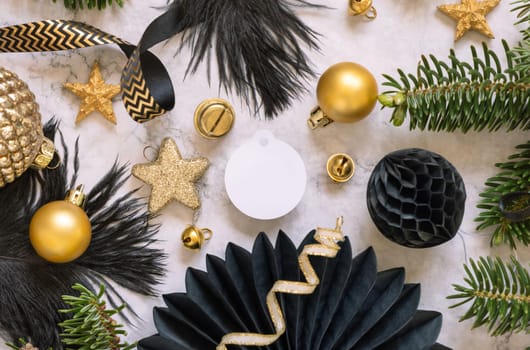 Paper round gift tag between fir tree branches and black and golden Christmas decorations on a white marble table top view. Mockup, copy space. Winter holiday flat lay with blank label