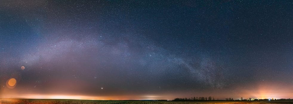 Night Starry Sky With Glowing Stars Above Countryside Landscape. Milky Way Galaxy And Rural Field Meadow In Early Spring. Panorama, Panoramic View.