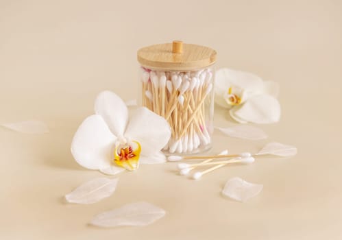 Bamboo cotton buds in a jar near white orchid flowers on light beige, close up. Organic skincare product for everyday beauty routine. Eco friendly Biodegradable cotton swabs..