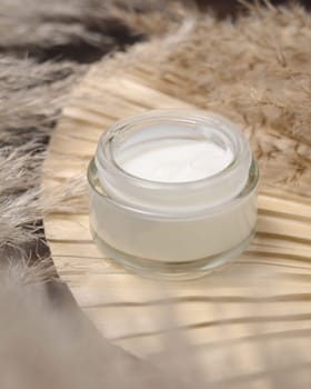 Opened cream jar on dried palm leaf near pampas grass close up, mockup. Skincare beauty product. Bohemian eco friendly cosmetics flat lay with dried leaves on wood.