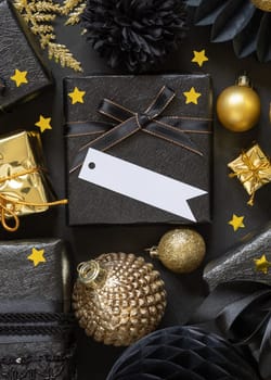 Black and golden Christmas Gift Boxes with a bow and ornaments close up, horizontal paper gift tag mockup, copy space. Dark winter composition with blank label card for Christmas, New Year, Birthday, Anniversary