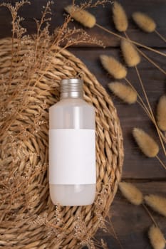 Cosmetic bottle on boho wattled placemat near Hare's Tail grass top view on wood, label mockup. Natural skincare product, cream or lotion. Bohemian eco friendly beauty flat lay.