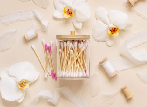 Bamboo cotton buds in a jar near white orchid flowers on light beige, top view. Organic skincare product for everyday beauty routine. Eco friendly Biodegradable cotton swabs..