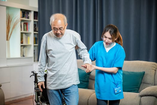 Asian doctor support old man to getting up to exercise, help handicapped elderly stand up, woman nurse assisting helping senior man patient get up from wheelchair for practice walking at home