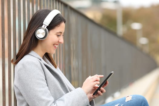 Side view portrait of a happy woman listening to music wearing headphones and using phone sitting in the street