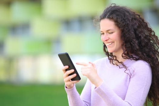 Happy woman with curly hair walks checking smart phone content in the street