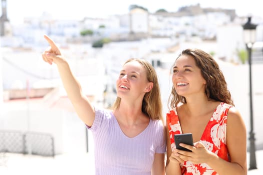 Two happy tourists pointing up holding smart phone in a white town on summer vacation