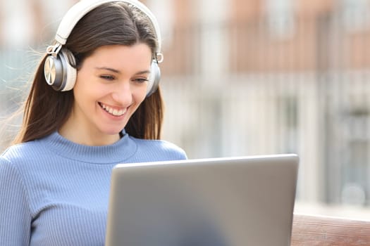 Happy woman using laptop with headphones sitting on a bench in the street