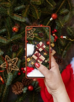 Woman's hand taking a mobile photo of christmas composition with Santa Claus hat, fir branches and holiday decorations top view. Blogger preparing content for social media using cell phone.