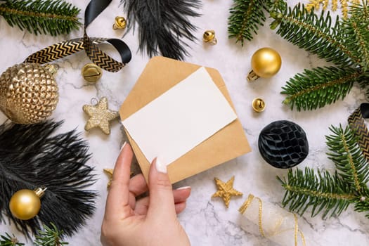 Hand holding Card and envelope, black and golden Christmas decorations and fir tree branches, top view. Winter flat lay with blank invitation card, Mockup, copy space