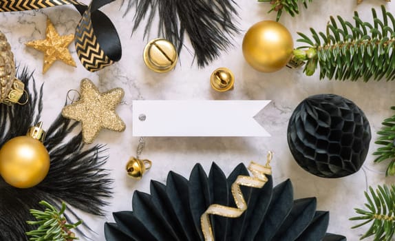 Paper horizontal gift tag between fir tree branches and black and golden Christmas decorations on a white marble table top view. Mockup, copy space. Winter holiday flat lay with blank label