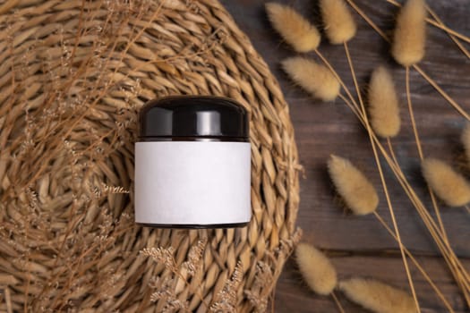 Cream jar on wattled boho placemat on wood near Hare's Tail grass top view, packaging label mockup. Natural skincare product for everyday beauty routine. Bohemian eco friendly cosmetics flat lay.