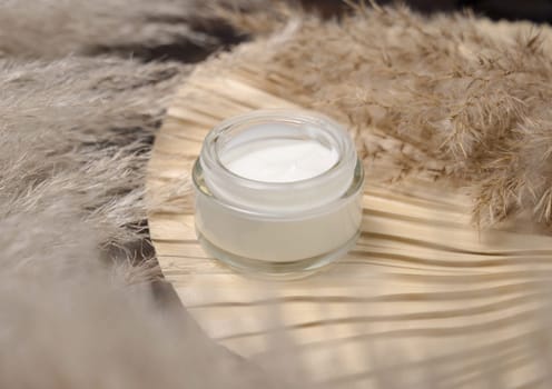 Opened cream jar on dried palm leaf near pampas grass close up, mockup. Skincare beauty product. Bohemian eco friendly cosmetics flat lay with dried leaves on wood.