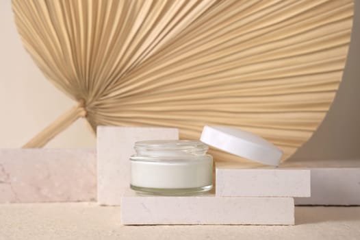 Opened glass cream jar with a white lid on travertine stone against dry beige palm leaf close up, mockup. Skincare beauty product - cream or lotion. Natural organic cosmetics. Eco friendly composition..