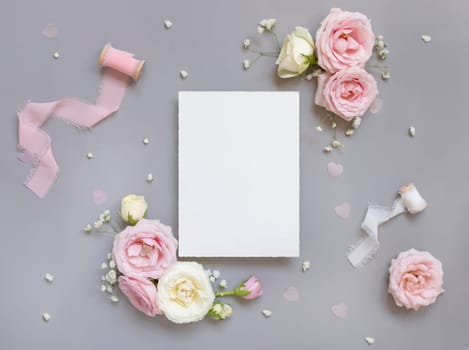 Paper card between light pink roses and silk ribbons on grey top view, wedding mockup. Romantic scene with blank vertical card and pastel flowers flat lay.. Valentines, Spring or Mothers day concept
