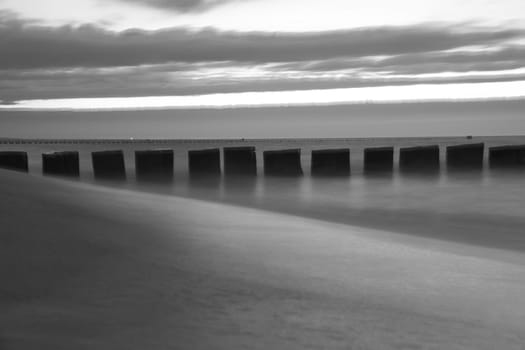 Sunset in black and white on the beach of the Baltic Sea. The groynes protrude into the sea. Blue hour with clouds in the sky. Landscape photo in Zingst