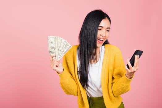 Portrait Asian happy beautiful young woman teen shopper smiling standing excited holding online smart mobile phone and dollar money banknotes on hand in summer, studio shot isolated on pink background