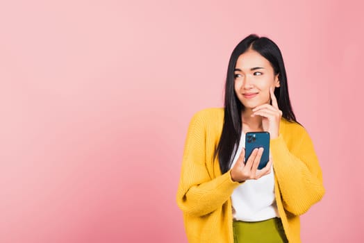 Happy Asian portrait beautiful cute young woman excited smiling holding mobile phone and think idea finger touch face, studio shot isolated on pink background, female using smartphone making gesture