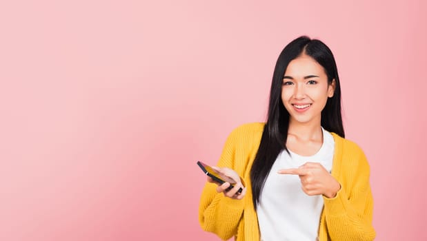 Portrait Asian happy beautiful young woman smile making finger pointing on smartphone in hand shopping online application looking to camera isolated, studio shot pink background with copy space