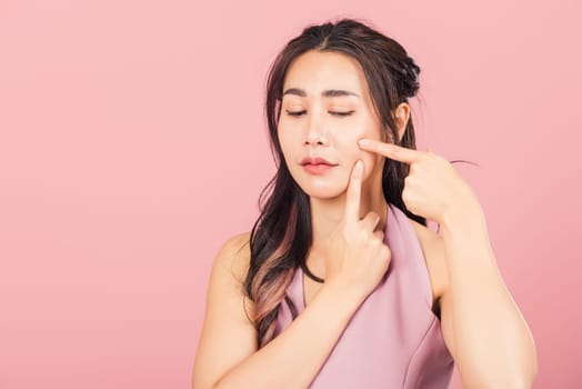 Portrait teenage Asian beautiful young woman having skin problems squeezing pimples on her face, studio shot on pink background, with copy space, Thai female acne, beauty care concept