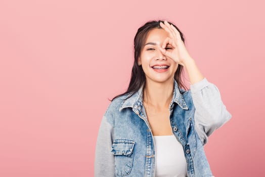 Portrait Asian beautiful young woman standing, She made finger OK symbol sign to agree near eyes looking at camera, studio shot isolated on pink background, Thai female agree hand sign copy space