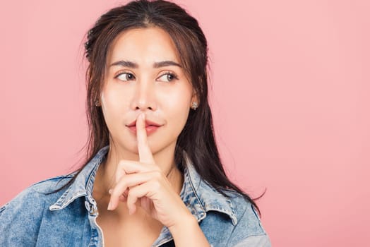 Portrait Asian beautiful young woman wear denim smile stand making finger on lips mouth silent quiet gesture, studio shot isolated on pink background, Thai female quiet hand sign copy space