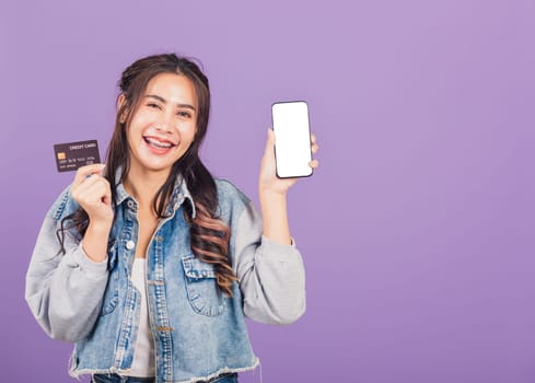Portrait of Asian beautiful young woman confident wear denim jeans shirt smiling show mobile phone blank screen and credit card bank, studio shot isolated on purple background