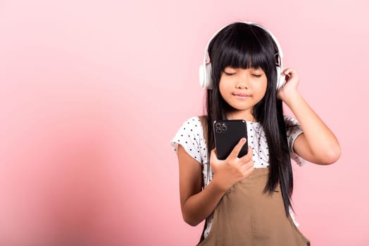 Asian kid 10 years enjoying listening music from mobile phone wear wireless headset closed eyes at studio shot isolated on pink background, Happy child girl use smartphone listen music with headphones