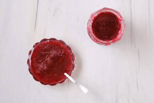 Top view of jar and saucer with cherry jam on white wooden background