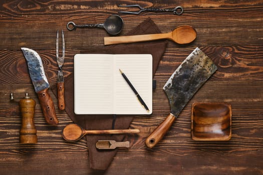 Top view of vintage steak fork, knife and hatchet on wooden table next to notebook. Barbecue appliances and open notepad, food blogging and menu concept.