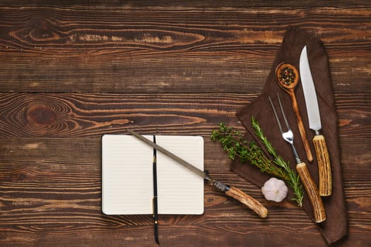 Vintage steak fork, knife and sharpener on wooden table next to notebook. Barbecue appliances and open notepad, food blogging and menu concept.