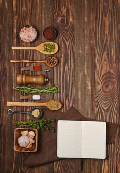 Top view of open notepad for recipes next to kitchen spoons and bowls with various spices, herbs and seasonings. Cooking concept of spicy and piquant food