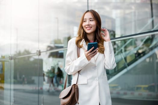 Portrait successful business woman smiling holding smartphone use application chat online in morning, Asian businesswoman working with mobile phone standing against street front building near office