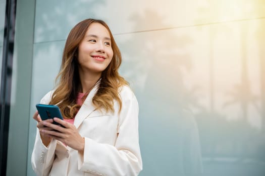 Portrait successful business woman smiling holding smartphone using app chat online in morning, Asian businesswoman typing text on mobile phone standing outdoors street front building near office