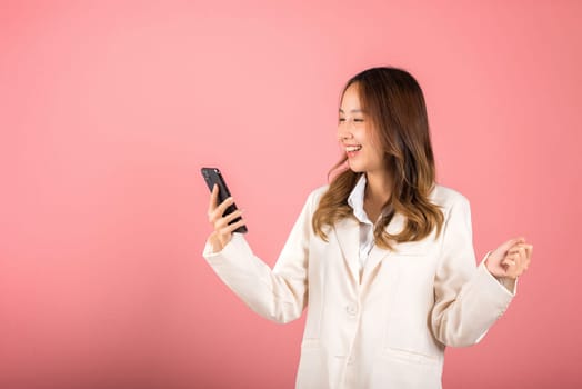 Happy Asian portrait beautiful cute young woman teen smiling excited face using mobile phone say yes! Studio shot isolated on pink background, Thai female surprised make winner gesture on smartphone